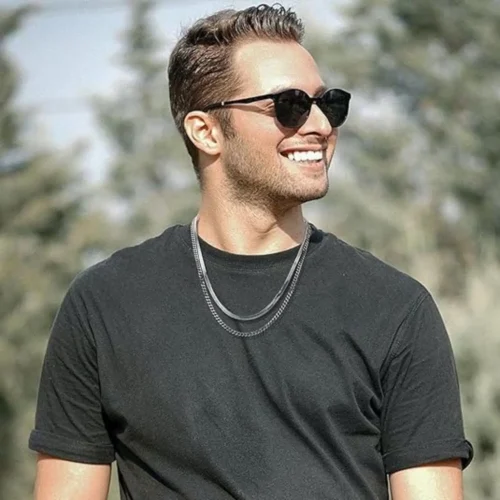 Hombre sonriente con gafas de sol y doble cadena plateada, look al aire libre.